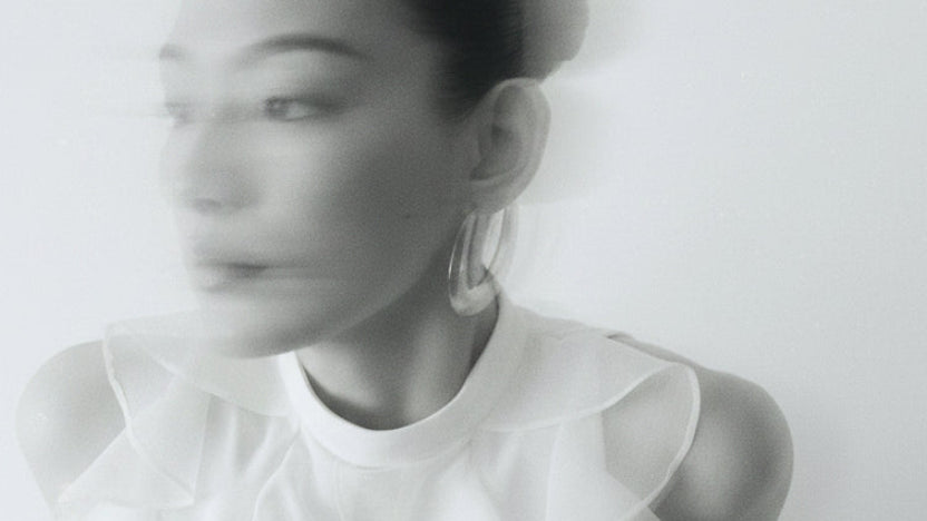 A woman wearing a white ruffled sleeveless blouse with a high mock neck and side cutout, paired with large gold hoop earrings, photographed against a plain light background.